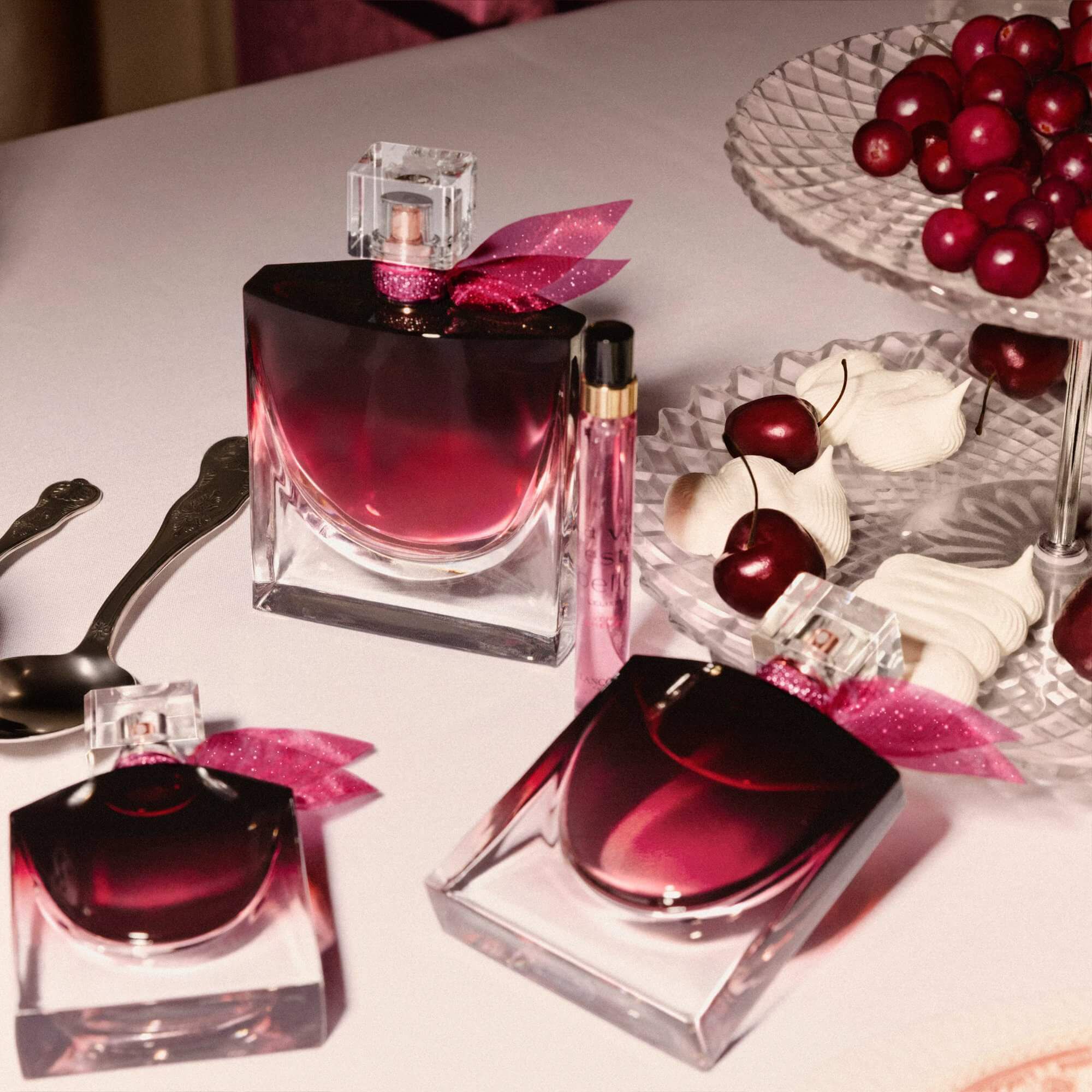 An elegant tabletop scene featuring multiple sizes of the perfume bottle alongside a bowl of fresh cherries and silver spoons.	