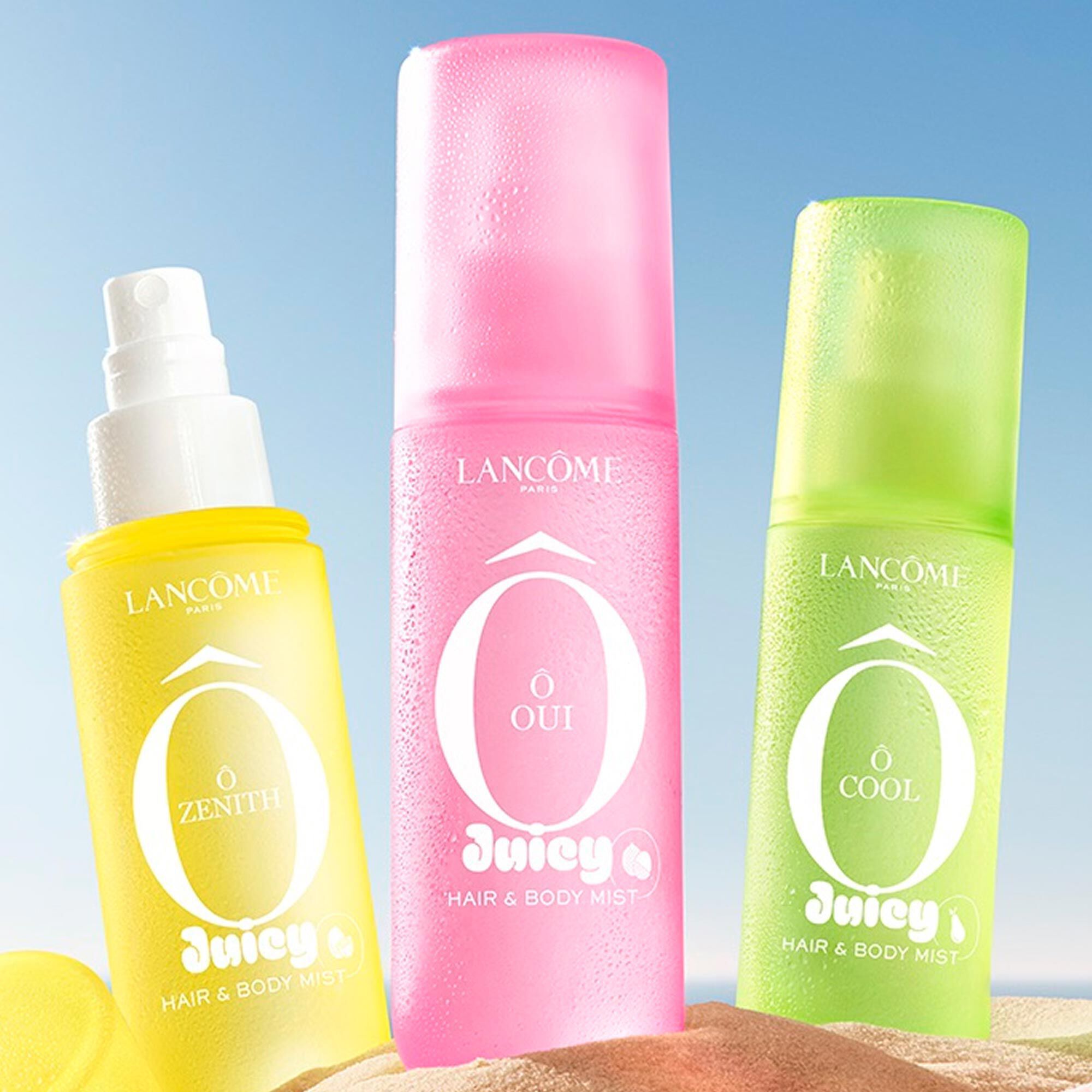 Three colorful spray bottles, one yellow, one pink, and one green, standing on a sandy beach next to a pink striped towel. The ocean is in the background under a clear blue sky. 