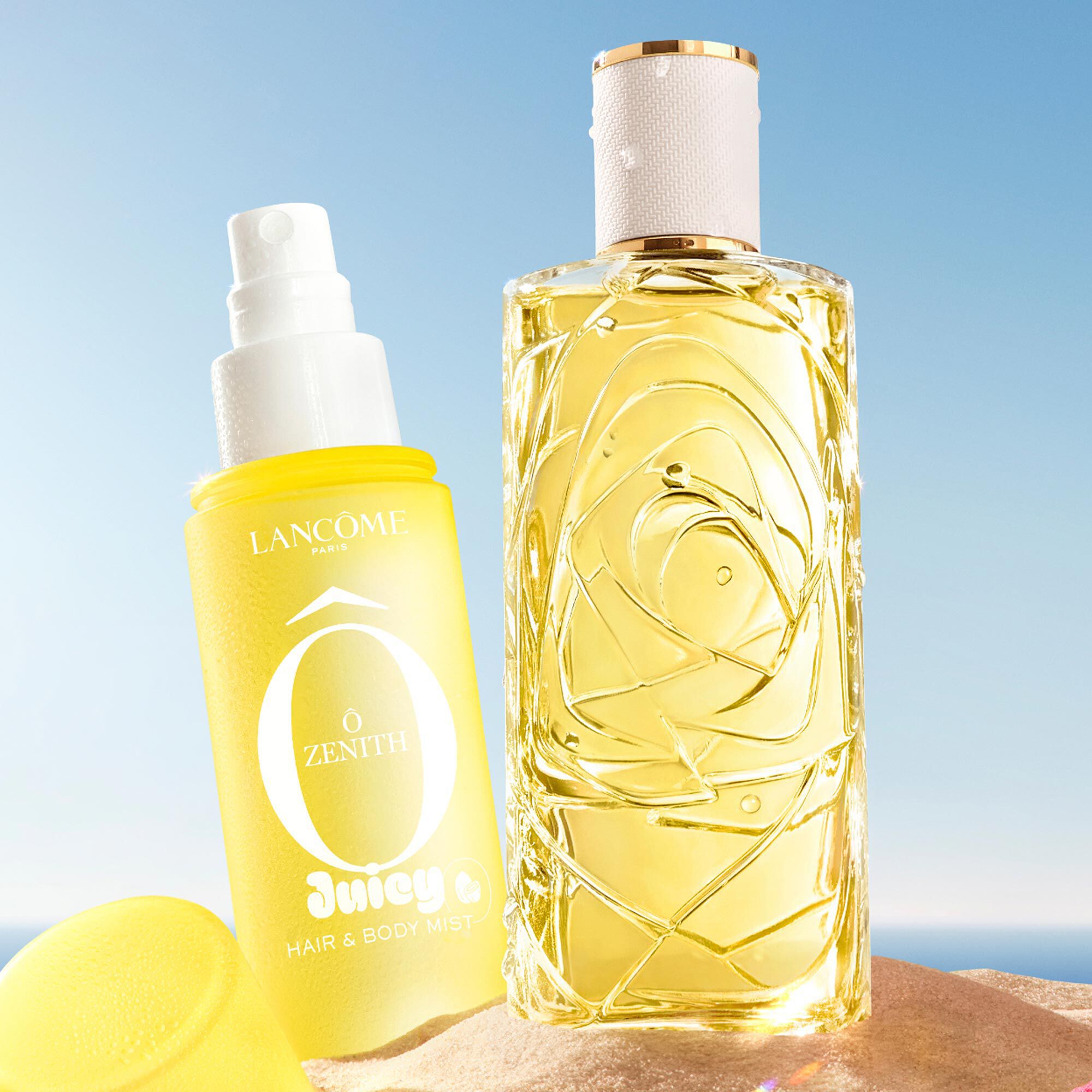 Two yellow bottles, one perfume rectangular bottle with a textured design and one spray bottle with a yellow cap on the side, standing on a sandy beach. A folded pink striped beach towel lies next to the bottles. In the background, there is a clear blue sky and the ocean.