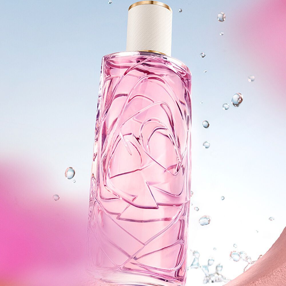 Textured light pink perfume bottle with water droplets, seen through a circular opening in a pink wall. Light blue sky background with blurred pink flowers in the foreground and background.