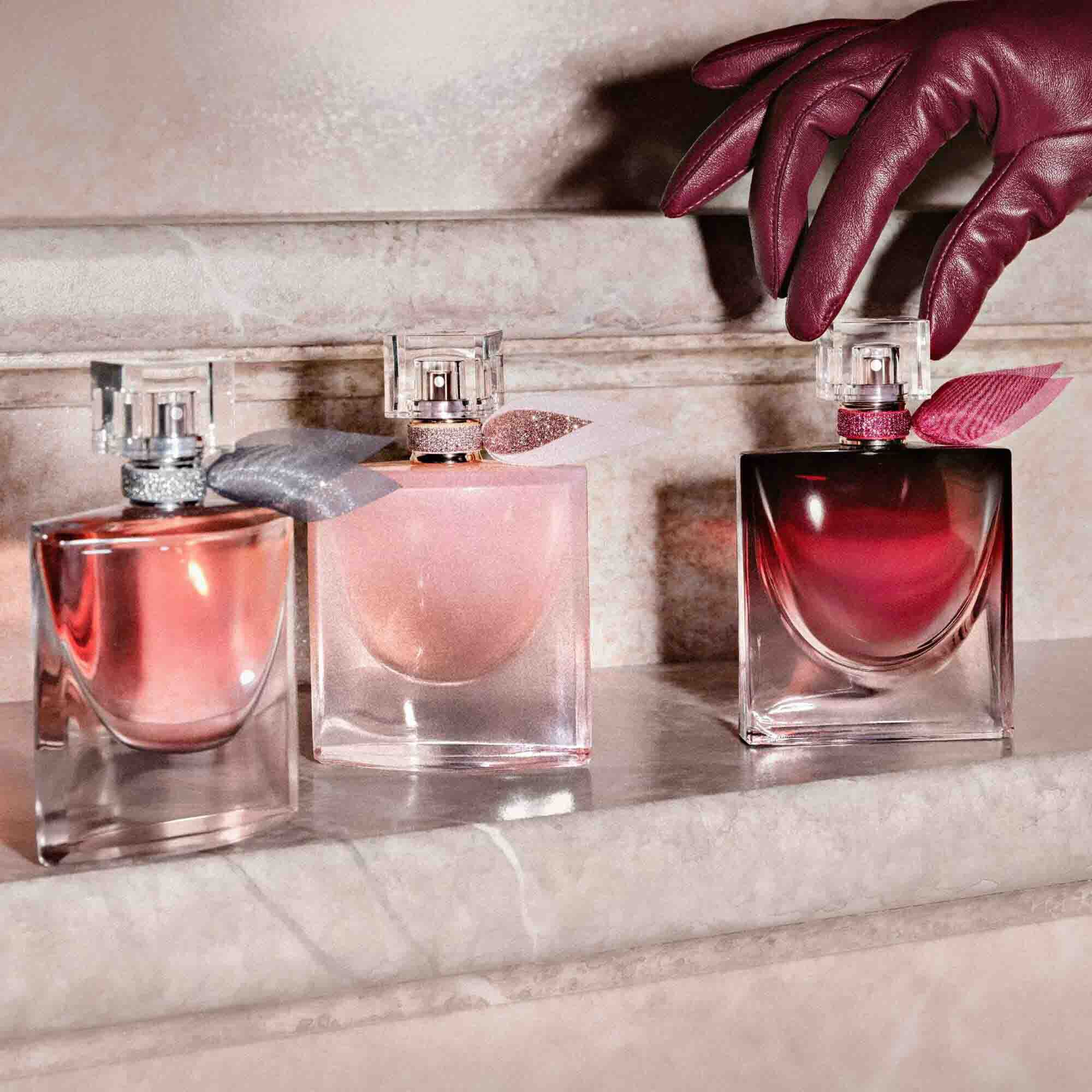 Three different versions of La Vie Est Belle in varying shades of pink and burgundy are lined up on a stone step being reached for by a hand in a leather glove.	