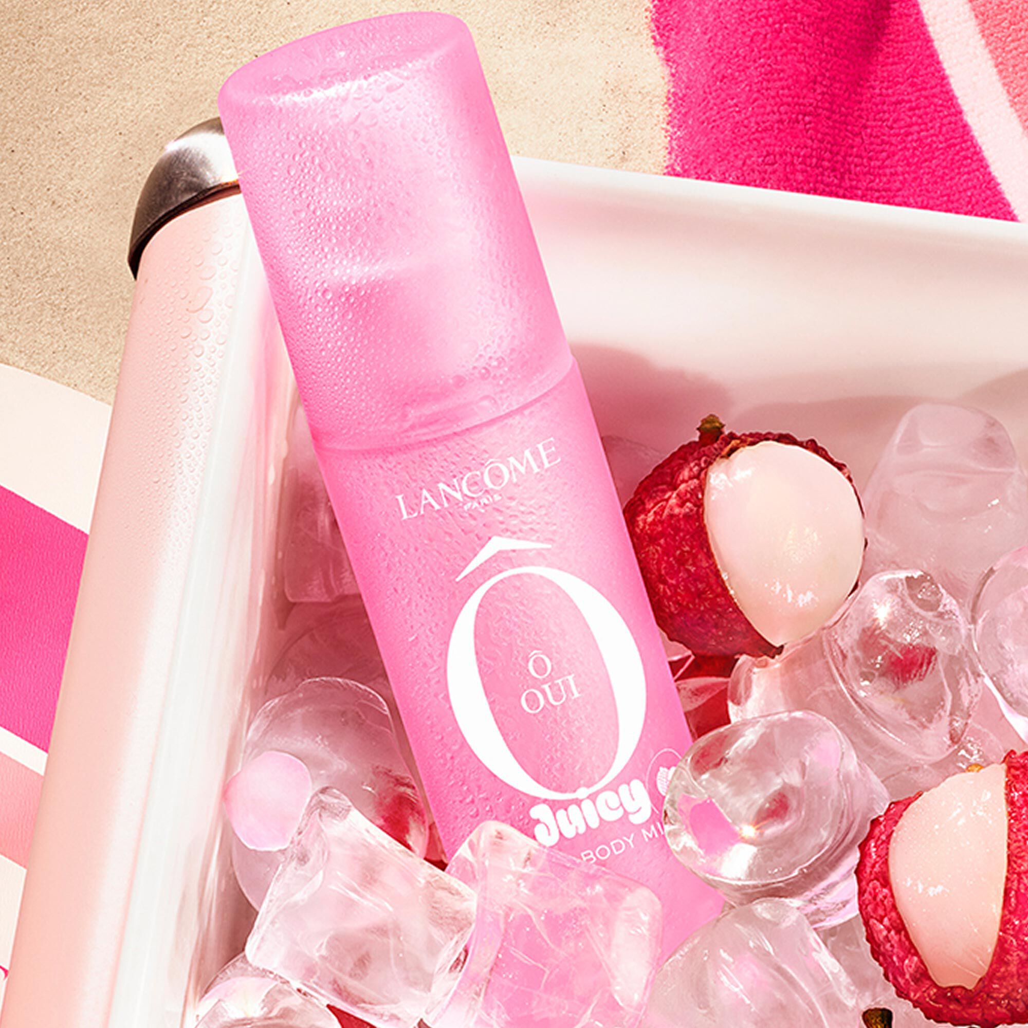 A pink mist bottle in a cooler filled with ice cubes and lychees. Next to it a pink paddle and a striped pink towel on a sandy background.